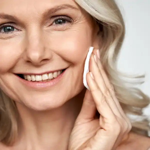 A close-up beauty portrait showing bright blue eyes, platinum blonde hair, and a warm smile while applying skincare.